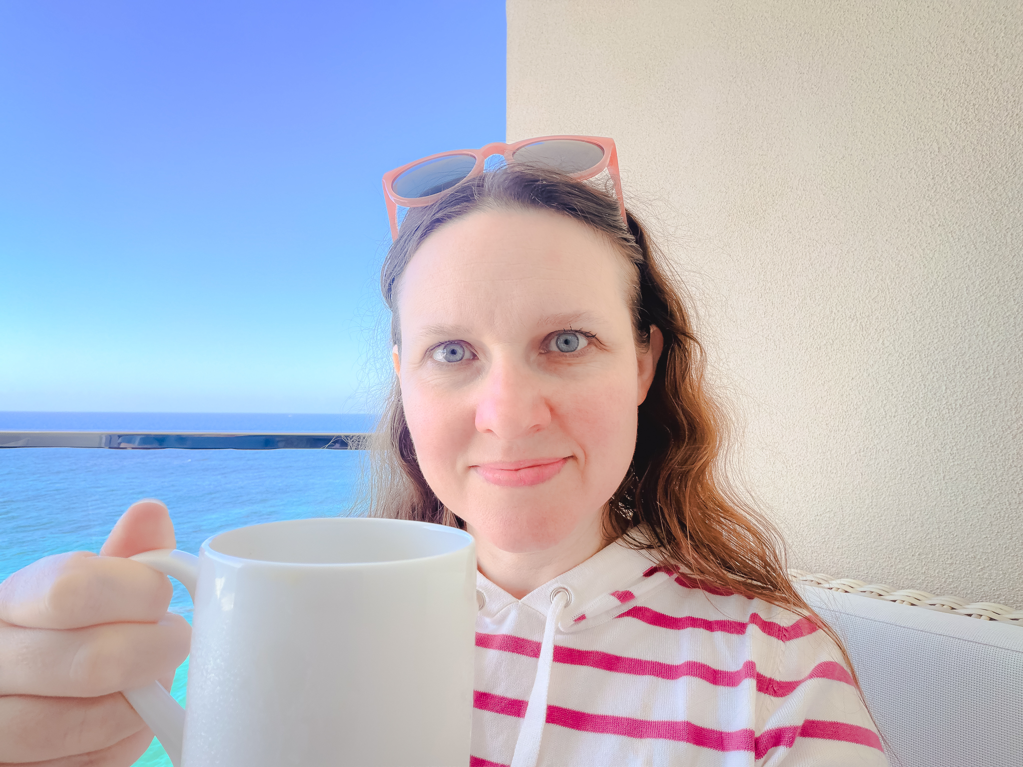 Shannon holding up a coffee cup with the ocean in the background. Shannon is a woman with fair skin, blue eyes, and light brown hair.